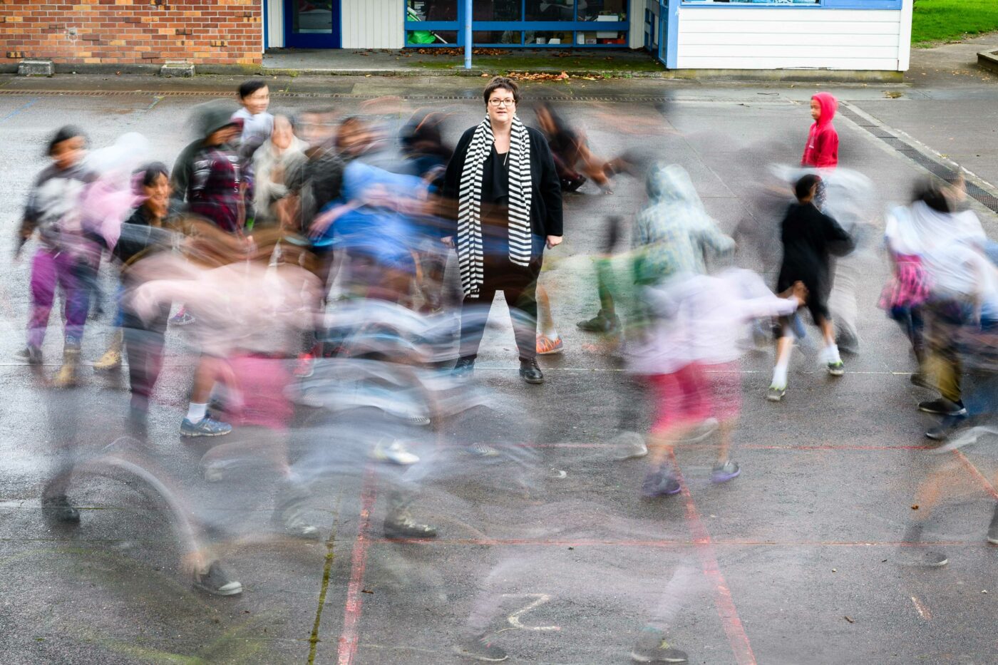A principal of a primary school standing in the centre of a courtyard with an action shot of children running in a circle around her.